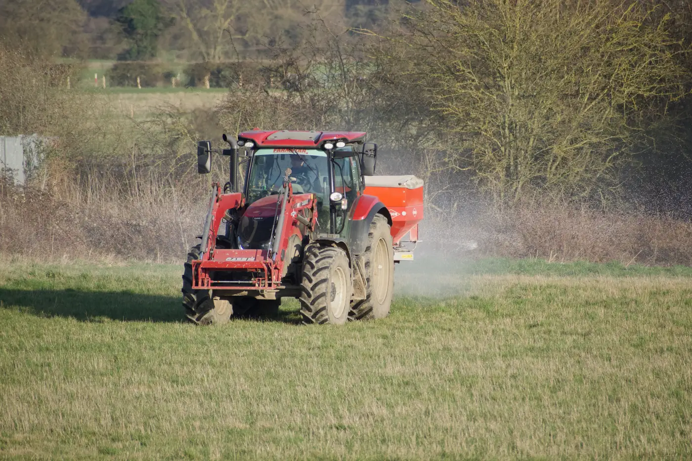 A red tractor pulling a dry fertilizer spreader across a farm field in daylight.