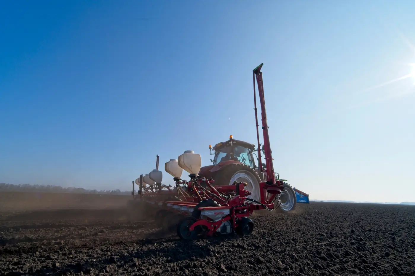 A tractor pulling a planter, sowing seed rows into dark tilled soil under a clear sky.